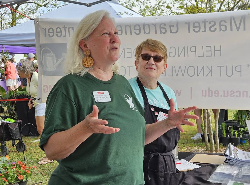 Two women stand at an outdoor event in front of a Master Gardener banner. The woman in green speaks, while the other in black listens.